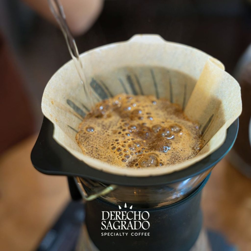 Close-up of freshly ground coffee brewing in a paper filter over a glass carafe at Derecho Sagrado, with water being poured from a kettle.