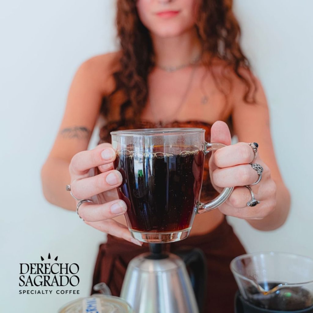 A woman holding a clear glass mug of black coffee, with other coffee preparation items in the background.