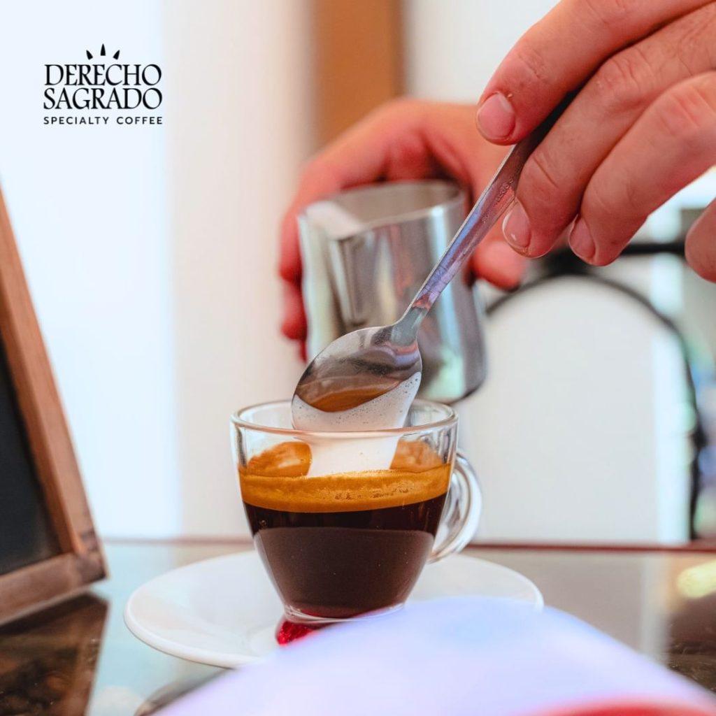 Close-up of a hand stirring a freshly brewed macchiato in a clear glass cup at Derecho Sagrado.